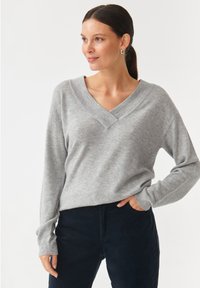Grijze V-halstrui van zacht materiaal, met lange mouwen en een ontspannen pasvorm. Gecombineerd met donkerblauwe broek.