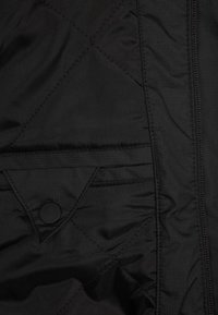 Svart vadderad jacka med en slät yta, med ett diagonalt mönster och en tryckknappsficka på framsidan. Dragkedjan går längs ena sidan.