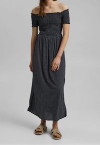 Abito maxi blu navy scollato sulle spalle con piccoli motivi geometrici bianchi, corpetto aderente e gonna fluida, abbinato a sandali beige.