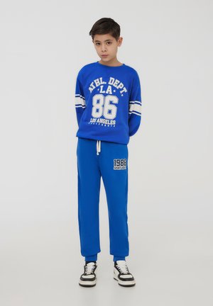 Felpa blu con grafica bianca "ATHL DEPT LA 86" e maniche a righe, abbinata a pantaloni tuta blu e sneaker bianco e nero.