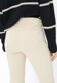 Lys beige jeans med et tætsiddende design og baglommer, båret sammen med en sort sweater med hvide striber. Blød tekstur, rene linjer.