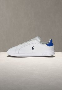 Λευκό χαμηλό αθλητικό παπούτσι με μπλε ταμπέλα στη φτέρνα, μαύρο λογότυπο Polo στο πλάι, λευκά κορδόνια και καθαρή ελαστική σόλα.