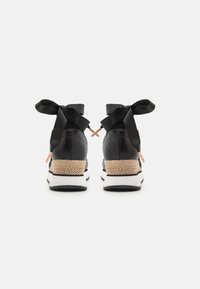 Black sneakers with a textured leather upper, featuring a woven jute heel, white midsole, and black satin ribbons tied at the ankle.