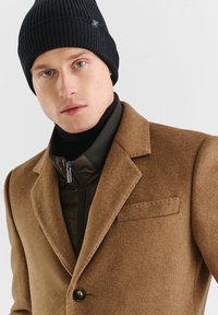 Brązowy płaszcz z mieszanki wełny z klapami i przednią kieszenią, założony na czarną pikowaną kurtkę z zamkiem. Czarna dziana czapka dopełnia całość.