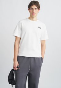 Λευκό βαμβακερό t-shirt με κοντά μανίκια, με μικρό μαύρο λογότυπο στο αριστερό στήθος, συνδυασμένο με γκρι παντελόνι φόρμας.