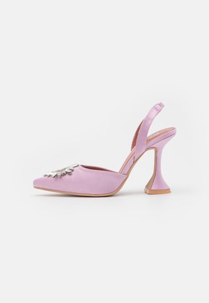 Zapato de tacón alto con tira en color lavanda, punta afilada, tacón acampanado y decoración ornamental plateada en la parte delantera sobre fondo blanco.