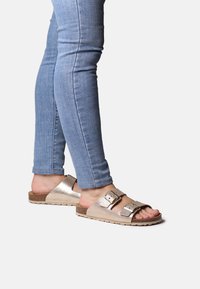 Tvåtone metalliska guldskor i sandalmodell med två justerbara remmar, korksålar och öppen tådesign, parat med ljusblå jeans.