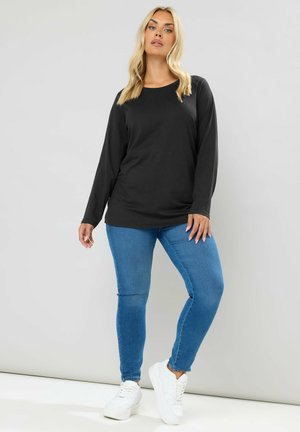Vrouw met blond haar die een zwart shirt met lange mouwen, blauwe spijkerbroek en witte sneakers draagt, staand tegen een grijze achtergrond.