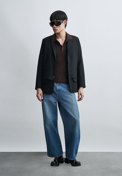 Άνδρας που φοράει μαύρο σακάκι, σκούρο καφέ πόλο πουκάμισο, φαρδιά μπλε τζιν παντελόνια, μαύρα μποτάκια και μεγάλα μαύρα γυαλιά ηλίου, στέκεται μπροστά σε μονόχρωμο φόντο.