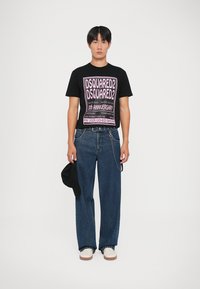 T-shirt graphique noir avec du texte rose, associé à un jean en denim bleu ample. Un chapeau noir est tenu dans une main. Des baskets blanches complètent la tenue.