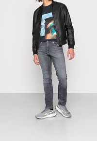 Svart läderbomberjacka, grafisk t-shirt med blå och orange design, ljusgrå jeans och grå sneakers med svarta detaljer.