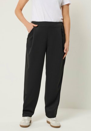 Person iført sorte viddebenbukser med sidelommer, hvide sneakers og en hvid kortærmet top, stående foran en ensfarvet baggrund.