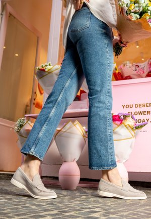 Person iført blå jeans og beige slip-on sko holder en buket og står nær indpakkede blomsterbuketter på en lyserød stand.