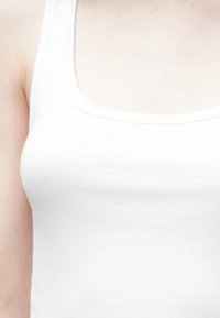 Λευκή ριμπ μπλούζα χωρίς μανίκια με φαρδιές τιράντες και στρογγυλεμένο λαιμόκοψη. Το ύφασμα φαίνεται μαλακό και ελαφρύ, με απαλό υφή.