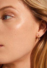 Gouden oorbel met een golvend ontwerp, vastgemaakt aan een doorboord oor. Gladde oppervlakte met een reflecterende afwerking, die huidtinten aanvult.