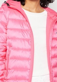 Różowa kurtka puchowa o błyszczącej powierzchni, z pikowanymi sekcją, z zamkiem błyskawicznym i kapturem; noszona na białym podkoszulku z prążkowanej tkaniny.