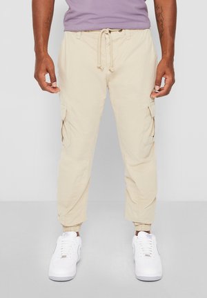 Mand iført beige cargobukser med snøre i taljen og hvide sneakers, stående foran en ensfarvet grå baggrund.