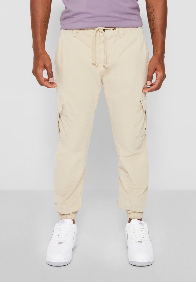 Homme portant un pantalon cargo beige avec cordon à la taille et des baskets blanches, debout devant un fond gris uni.