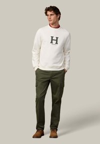 Witte sweatshirt met een "H" logo en "HACKETT" eronder, gecombineerd met olijfgroene cargobroek en bruine suède schoenen. Eenvoudig, casual ontwerp.