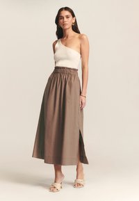 Falda marrón de cintura alta con cinturilla elástica y abertura lateral, combinada con una blusa blanca de un solo hombro de canalé y sandalias trenzadas.