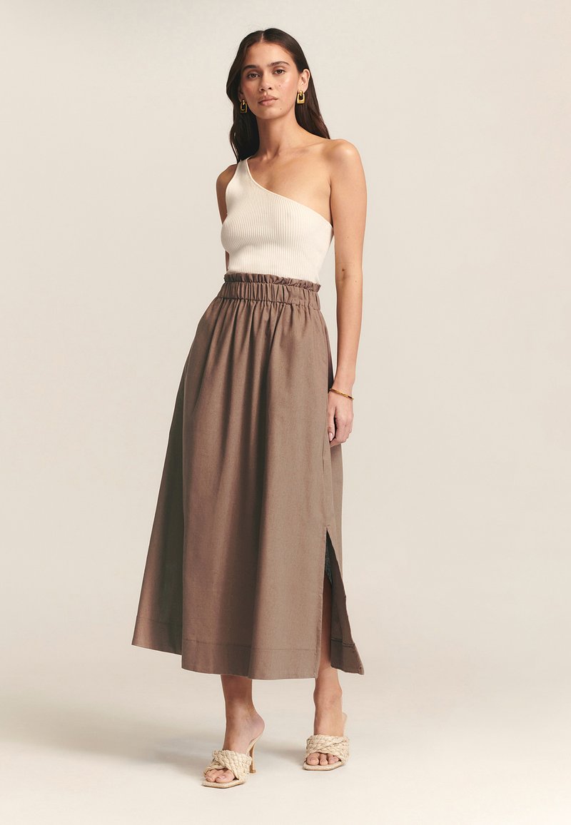 Falda marrón de cintura alta con cinturilla elástica y abertura lateral, combinada con una blusa blanca de un solo hombro de canalé y sandalias trenzadas.