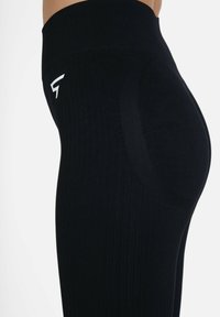 Črne rebraste legice z visokim pasom, ki imajo brezšivno zasnovo in majhen bel logo na bokih. Prilagodljiv, elastičen material.