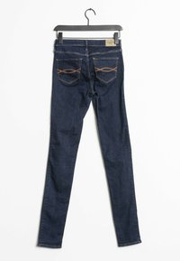 Úzké džíny v tmavě modrém džínovém materiálu, se středním pasem, zadními kapsami s oranžovým prošíváním a hladkým povrchem. Visí na černém ramínku.