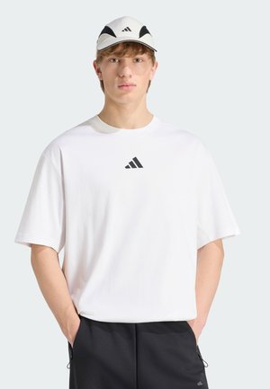 Jeune homme portant une casquette blanche Adidas et un T-shirt blanc oversize avec un logo noir Adidas, les mains dans les poches d’un pantalon noir, face à l’avant.