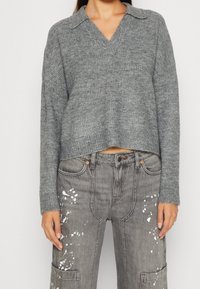 Grå ribbade tröja med v-ringning och långa ärmar, i kombination med grå denimbyxor med vit målarfärgssplatter och en djup framficka.