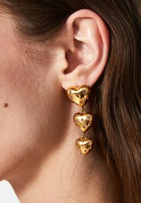 Pendientes en forma de corazón en tono dorado, con tres corazones apilados y adornados con pequeñas piedras de colores, que exhiben una textura suave y reflectante.