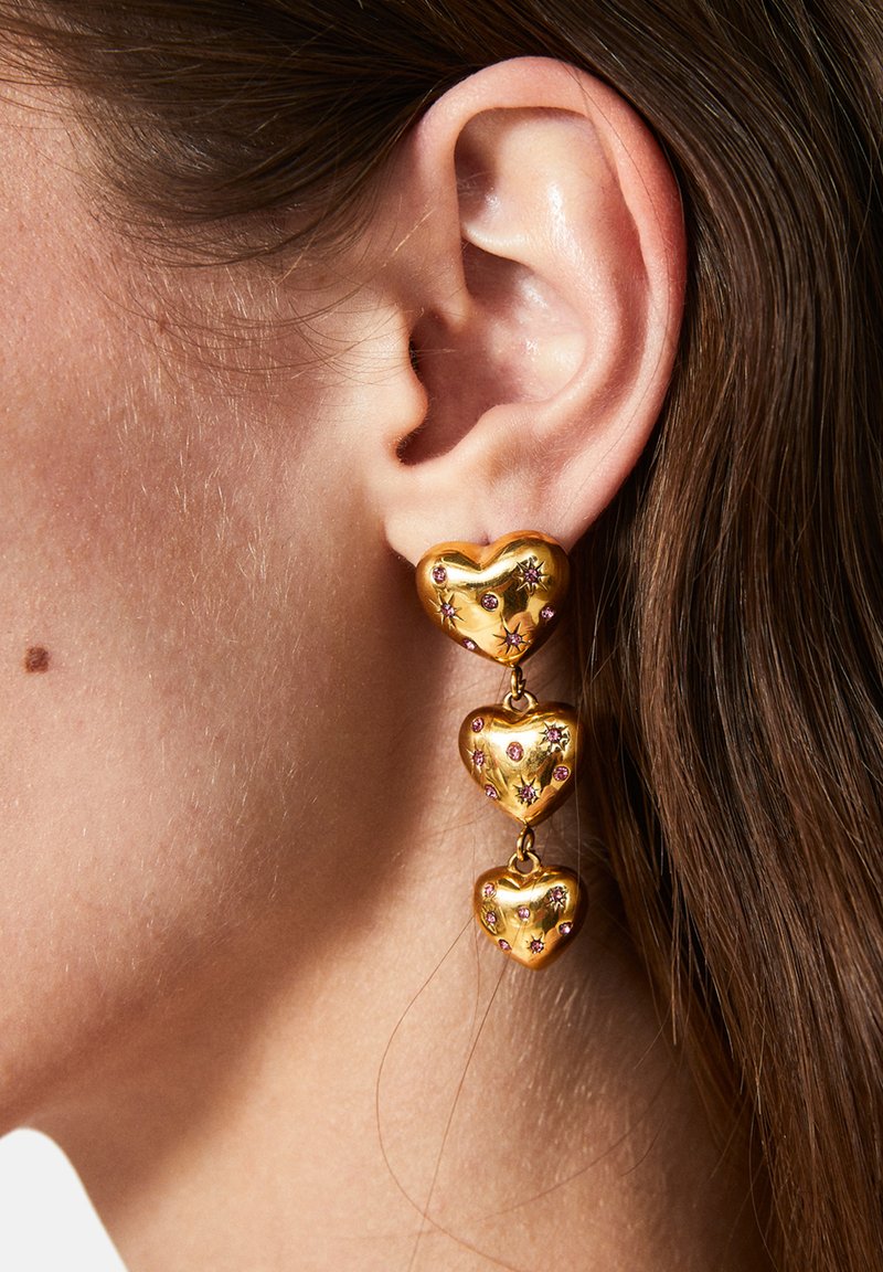Pendientes en forma de corazón en tono dorado, con tres corazones apilados y adornados con pequeñas piedras de colores, que exhiben una textura suave y reflectante.