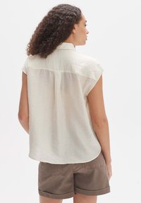 Hellbeige Kurzarmhemd aus strukturiertem Stoff mit feinen Falten auf der Rückseite und abgerundetem Saum, kombiniert mit braunen, hochgerollten Shorts.