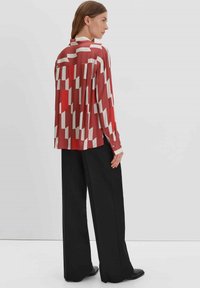 Langarmshirt in het rood met een geometrisch patroon in wit en crème. Soepele stof, met knoopschoenen en een ontspannen silhouet. Gemaakt in combinatie met zwarte broeken.