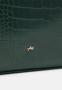 Pelle in pelle di coccodrillo di un verde scuro con accentuazione del logo in tonalità dorata. Finitura liscia, forma rettangolare, che mette in evidenza il caratteristico motivo rettiliano.