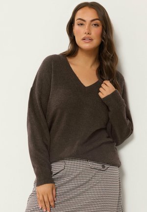 Femme aux longs cheveux foncés portant un pull marron foncé à col en V et une jupe à motif pied-de-poule, debout devant un fond clair.