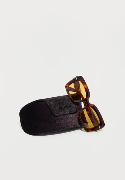 Ορθογώνια γυαλιά ηλίου με σχέδιο χελώνας και κεχριμπαρένιους φακούς, ακουμπισμένα σε μαύρη βελούδινη θήκη Tom Ford σε λευκό φόντο.