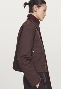 Brązowa krótka kurtka z dwoma przednimi kieszeniami i srebrnymi detalami, zestawiona z ciemnoczerwoną żeberkową szalem. Gładka tekstura, casualowy styl.