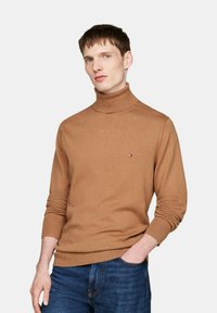 Brauner Rollkragenpullover aus weichem Stoff mit geripptem Kragen und Bündchen. Kleines Logo-Emblem auf der linken Brust. Getragen mit blauen Jeans.