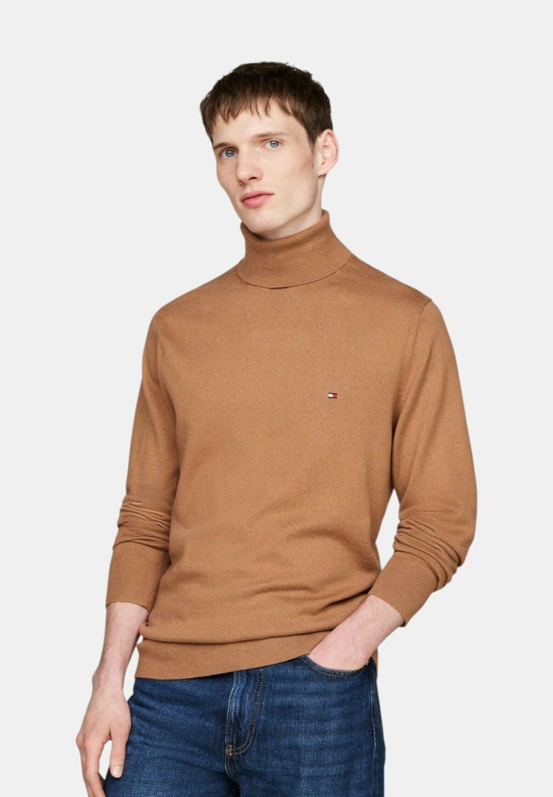 Brauner Rollkragenpullover aus weichem Stoff mit geripptem Kragen und Bündchen. Kleines Logo-Emblem auf der linken Brust. Getragen mit blauen Jeans.