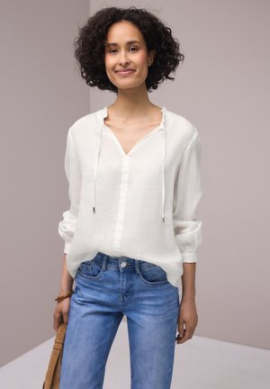 Femme aux cheveux bouclés portant un chemisier blanc et un jean bleu, debout et tenant un sac à main beige sur un fond neutre.