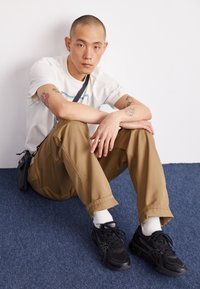 Baskets noires avec des motifs texturés, pantalon kaki ample et léger, et un t-shirt blanc avec un design graphique bleu, assis sur un tapis bleu.
