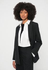 Kvinne med naturlig krøllete hår iført en svart blazer, hvit skjorte med knapper og svarte bukser, stående mot en ensfarget hvit bakgrunn.