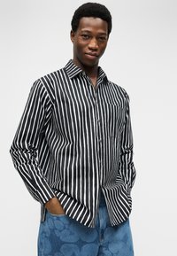 Νέος άνδρας που φοράει μαύρο και λευκό πουκάμισο με κάθετες ρίγες και τζιν με μπλε σχέδια, με τα χέρια στις τσέπες, μπροστά σε ανοιχτόχρωμο φόντο.