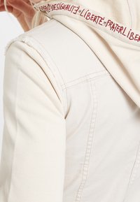 Persona vestida con una chaqueta de mezclilla color hueso, con la costura del hombro deshilachada y una capucha bordada en rojo con el texto "Liberté, Fraternité, Egalité".