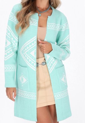 Femme portant un cardigan bleu clair à motifs sur une robe beige, accessoirisée d’un collier turquoise et d’une ceinture argentée avec des détails turquoise.