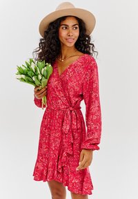 Červené květinové zavinovací šaty s dlouhými rukávy, volánkovým lemem a mašlí v pase. Modelka drží kytici bílých tulipánů.