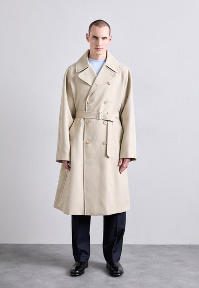 Beige Doppelreiher-Trenchcoat mit breitem Kragen, tailliertem Gürtel und seitlichen Taschen, kombiniert mit dunkelblauen Hosen und schwarzen Schuhen.