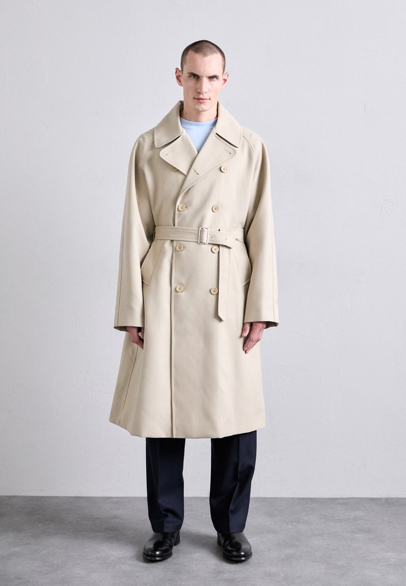 Trench coat beige doppiopetto con ampio colletto, vita cintrata con cintura e tasche laterali, abbinato a pantaloni blu navy e scarpe nere.