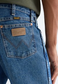 Close-up af en person iført blå Wrangler-jeans med læderlogo-mærkat på baglommen og grøn skjorte stoppet ind.