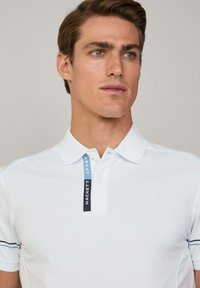 Polo bianco con colletto a marchio blu e nero, dotato di maniche corte e sottili righe orizzontali ai polsini.